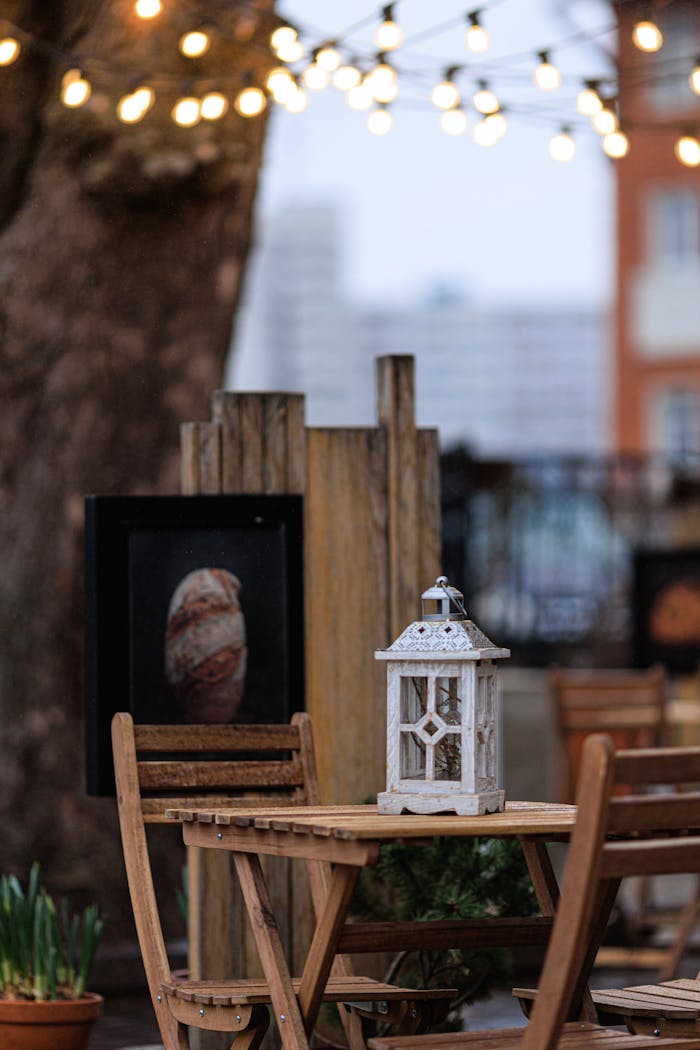 services-04 Charming outdoor patio with a rustic wooden table, chairs, and a decorative lantern under string lights.