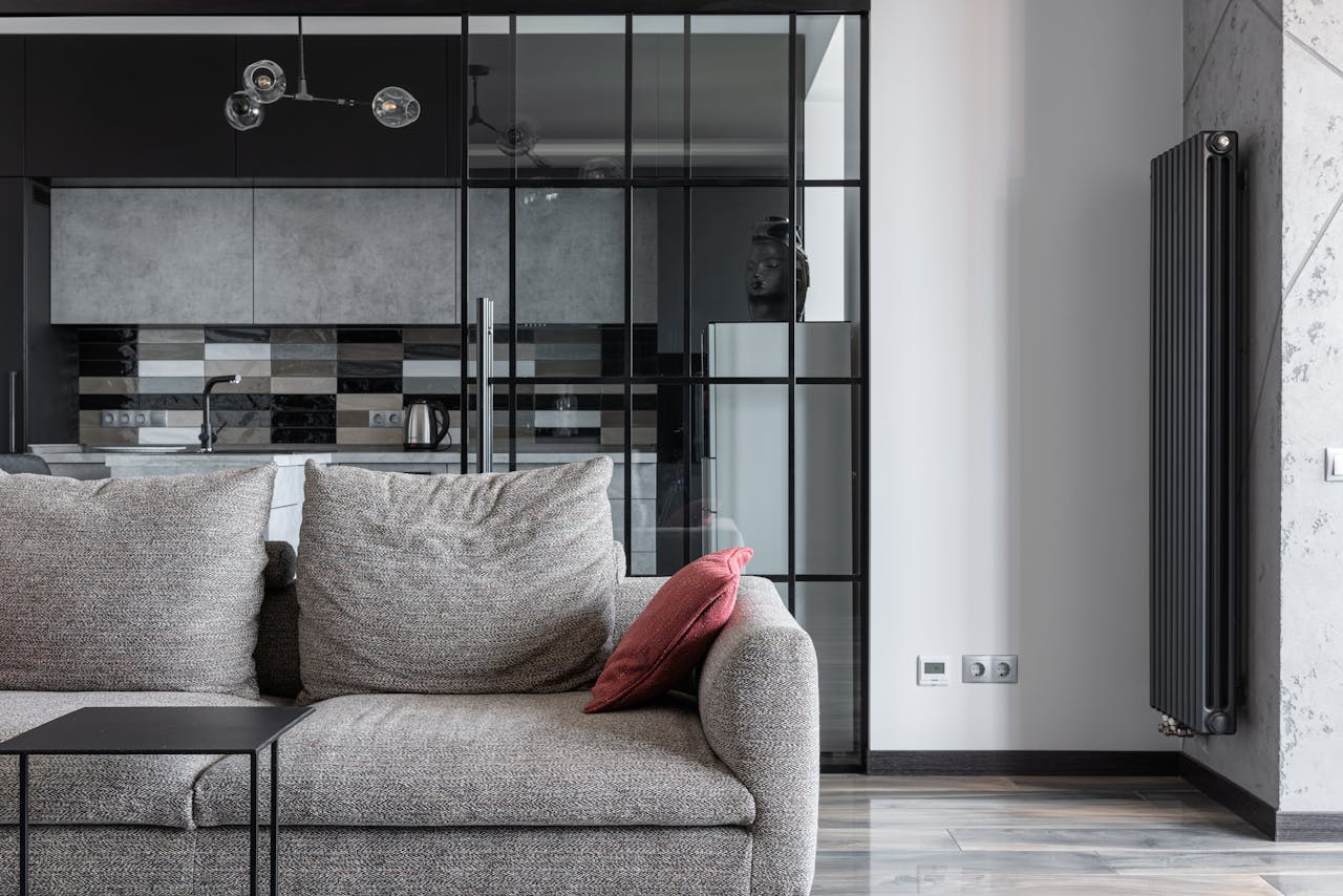 who-we-are Contemporary living space featuring a stylish gray sofa, red cushion, and a view into a modern kitchen.